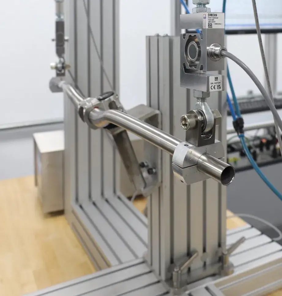 Industrial lab rig: a horizontal stainless-steel tube clamped to a vertical aluminum frame with sensors and fittings, cables in the background.